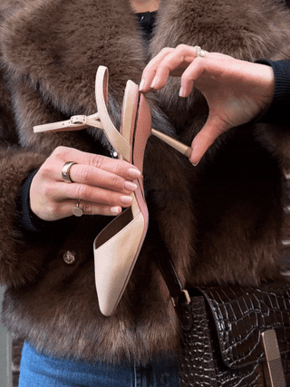 Person holding beige high-heeled shoe with ankle strap, wearing rings and brown fur coat