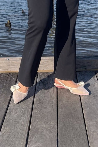 Feet wearing white mesh slingback flats with bows, standing on wooden dock by water