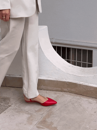 Red pointed-toe slingback shoe worn with cream pants and blazer, standing on tiled floor near white curved wall