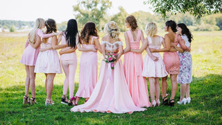 Bride and bridesmaids in pink dresses standing arm in arm outdoors, viewed from behind