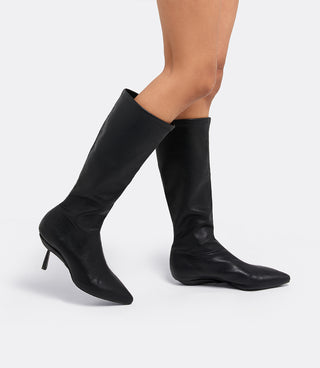 Model walking in the VICE VERSA Knee High Boots in black faux leather, one in heel mode and one folded flat