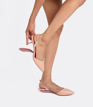 A model wearing light pink slingback pointed-toe flats, shown from the waist down as they adjust the ankle strap of one shoe against a light gray studio background.