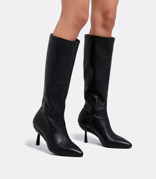 Close-up of a model wearing the VICE VERSA Knee High Boots in black faux leather with pointed toes and slim convertible heels, side view