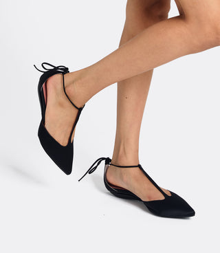 A pair of black pointed-toe flat slingback shoes with thin ankle ties, shown on a model's feet against a light gray studio background.