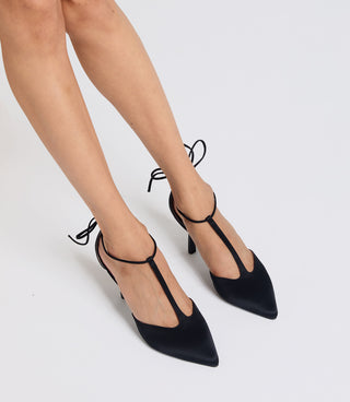 Black pointed-toe slingback heels with T-strap and ankle tie details, shown on a model's feet against a light gray studio background.
