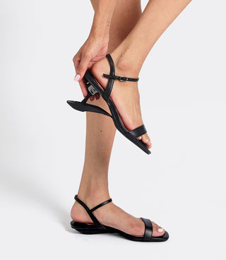 A person wearing black minimalist flat sandals with ankle straps, shown against a light gray studio background. One foot is flat on the ground and the other is lifted by a hand to show the side profile of the sandal.